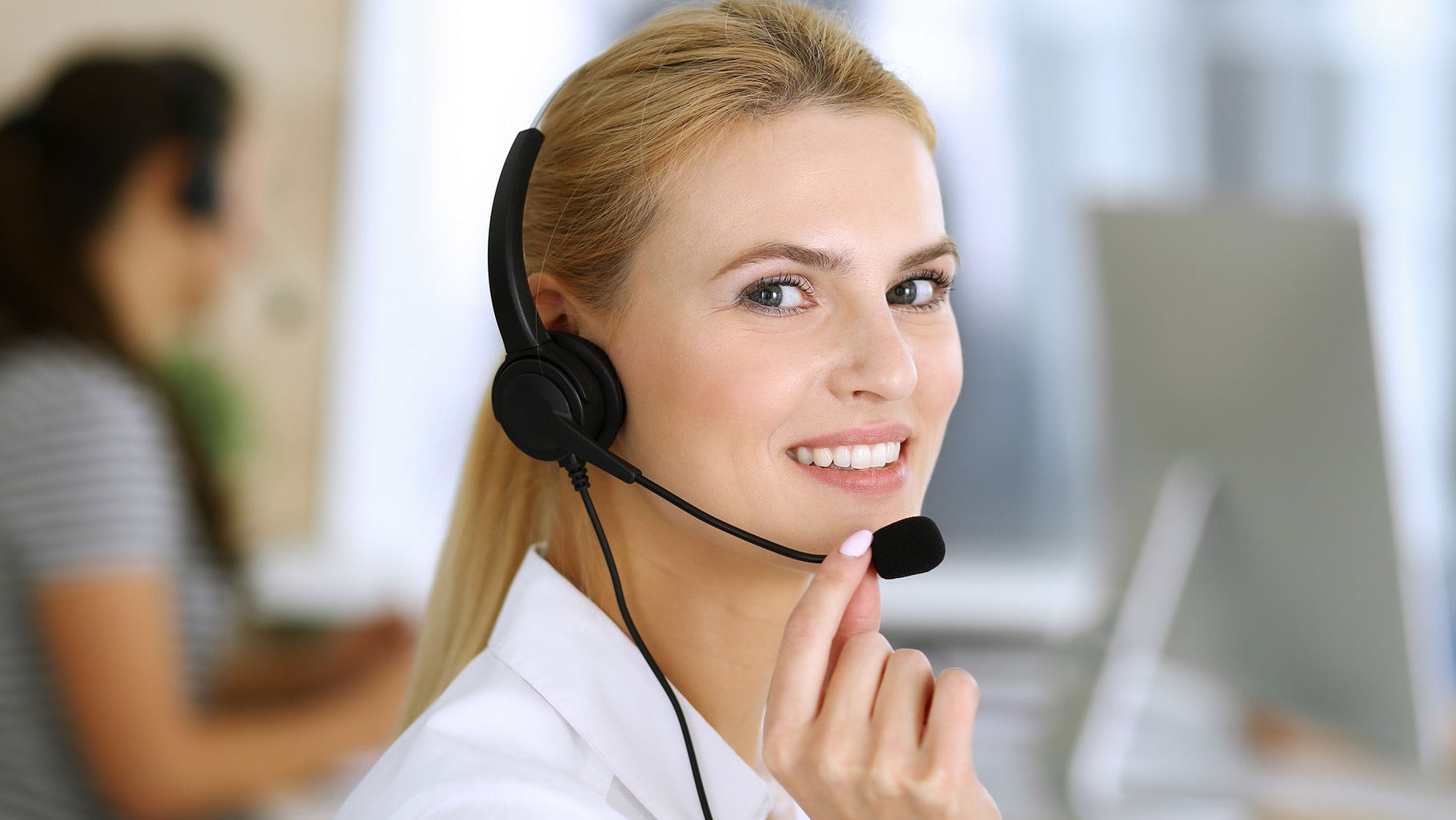 Blonde business woman using headset for communication and consulting people at customer service office. Call center. Group of operators at work at the background
